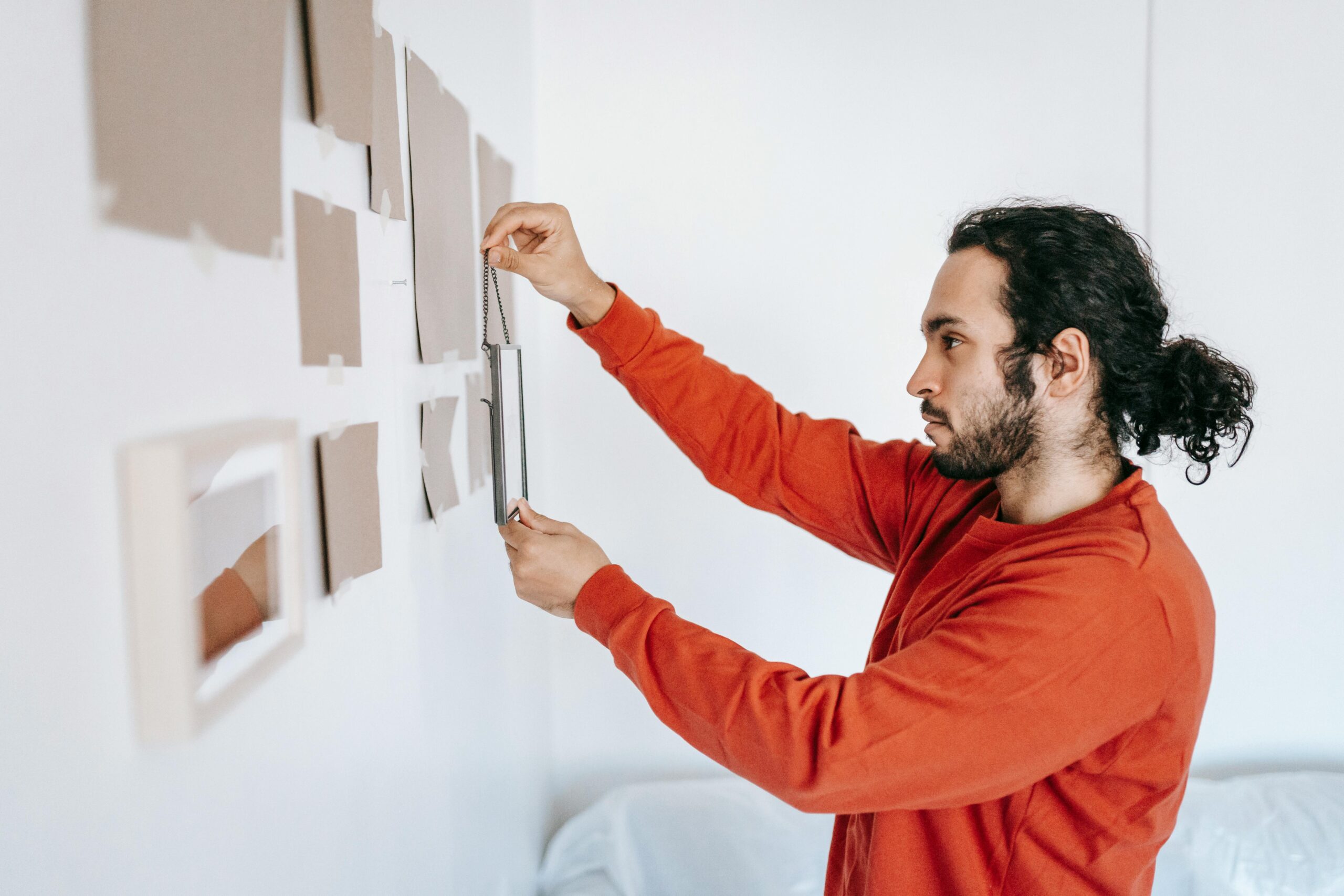 Young man in red sweater carefully arranging picture frames on a wall in a bright room.